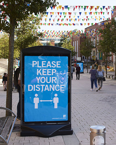 A photograph of the University precinct, including a large 'Please keep your distance' sign